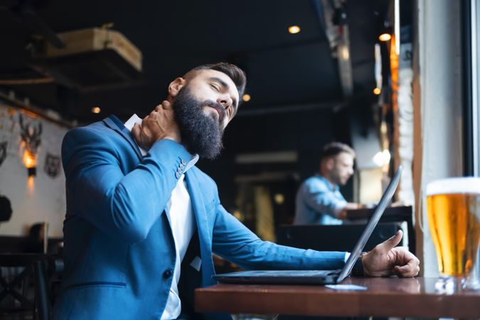 Man stretching neck while working on his laptop in a bar