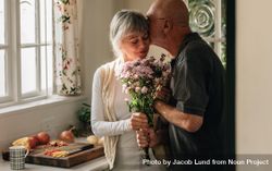 Older couple standing in kitchen holding a bunch of flowers 5qDYj0