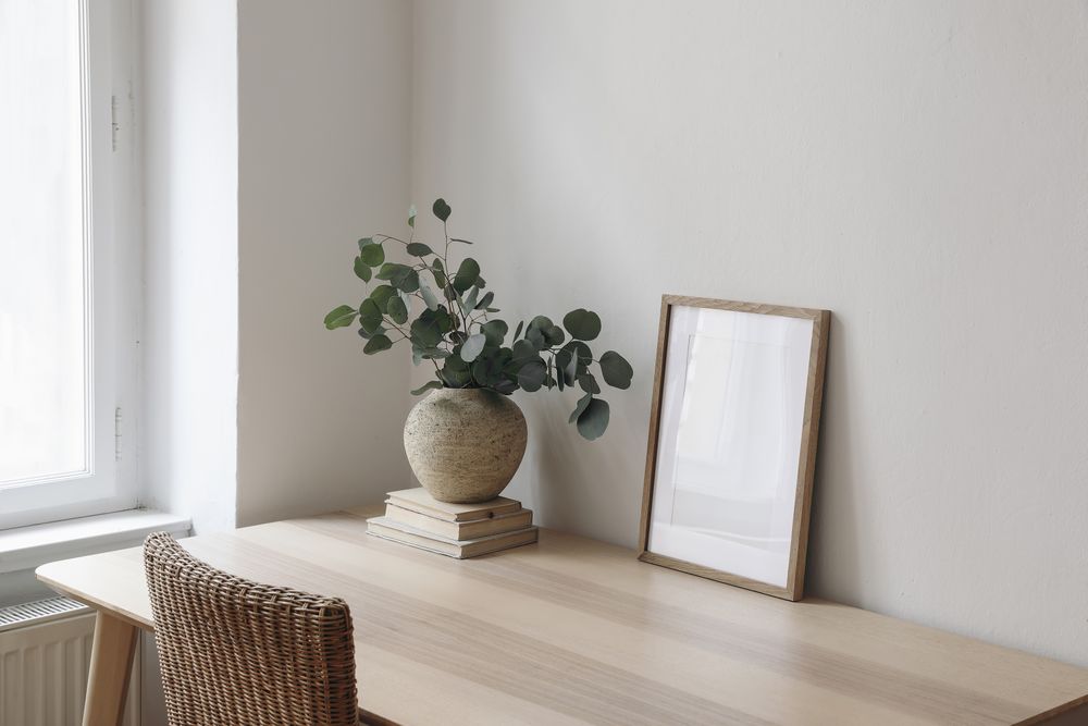 Living room with vase, eucalyptus tree branches, old books on wooden table on rattan chair near window with mockup poster