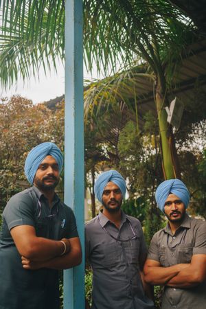 Three Indian men in blue turbans and gray shirts posing under palm tree in Dera Baba Nanka, India