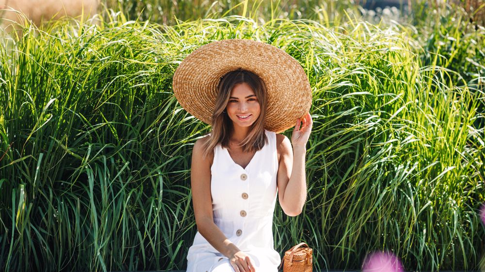 Happy woman sitting in front of long grass with straw hat