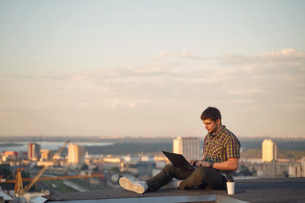 Man sitting on roof over looking city working on laptop with takeaway drink