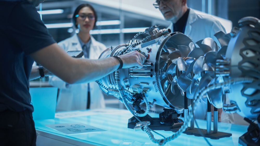 Diverse Multiethnic Team of Industrial Engineers Gathered Around a Table With a Prototype Turbofan Engine.