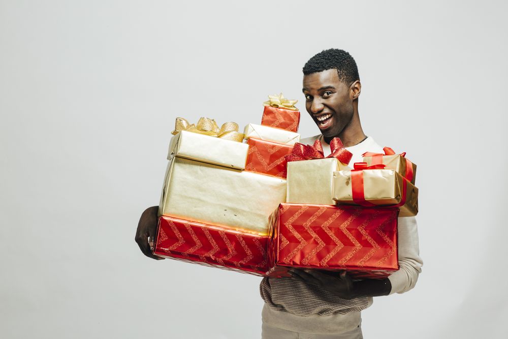 Black man making funny face holding pile of wrapped presents in both arms as glitter falls
