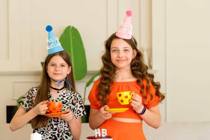 Two girls wearing birthday hats and holding colorful cups
