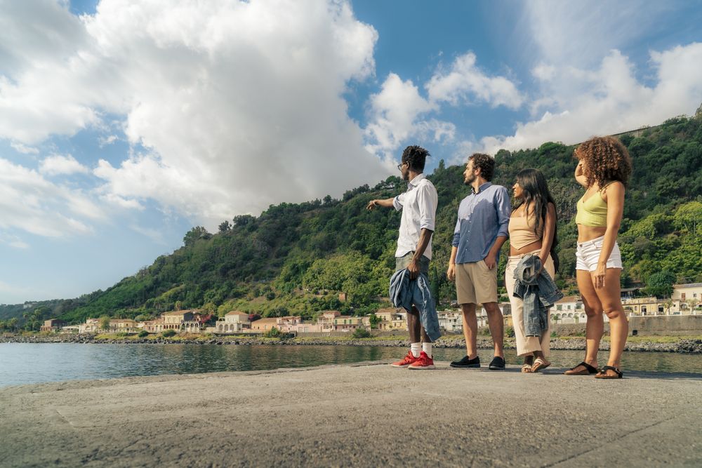 Group of diverse friends on a pier, with one man pointing and looking at the coastal view.