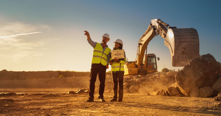 White Male Civil Engineer And Hispanic Female Architect Walking On Construction Site With Laptop Computer And Talking About New Real Estate Project. Machinery Working On Background. Golden Hour.