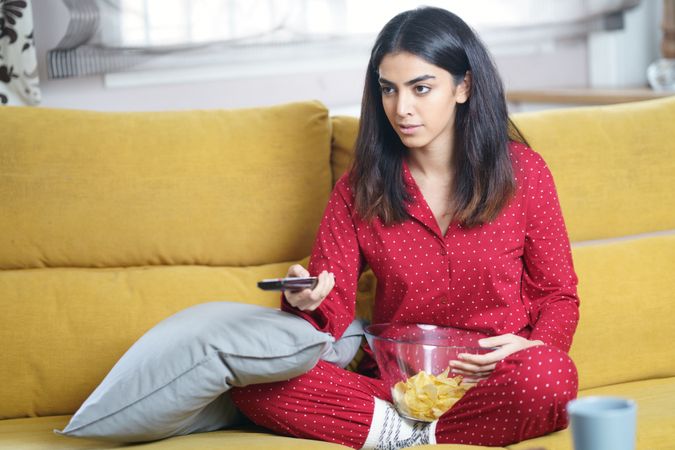 Woman in red pajamas sitting on sofa with bowl of chips and remote control