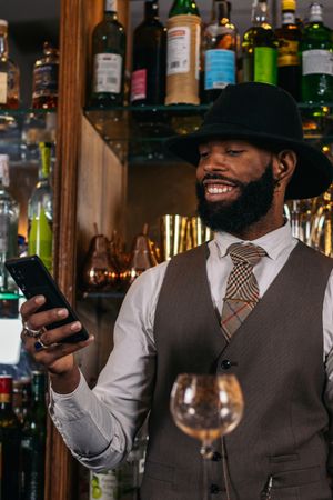 Stylish Black bartender in hat using smartphone while making cocktail at cocktail bar