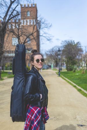 Side view of a young smiling hipster woman walking in a park in sunny day while carrying a guitar on back