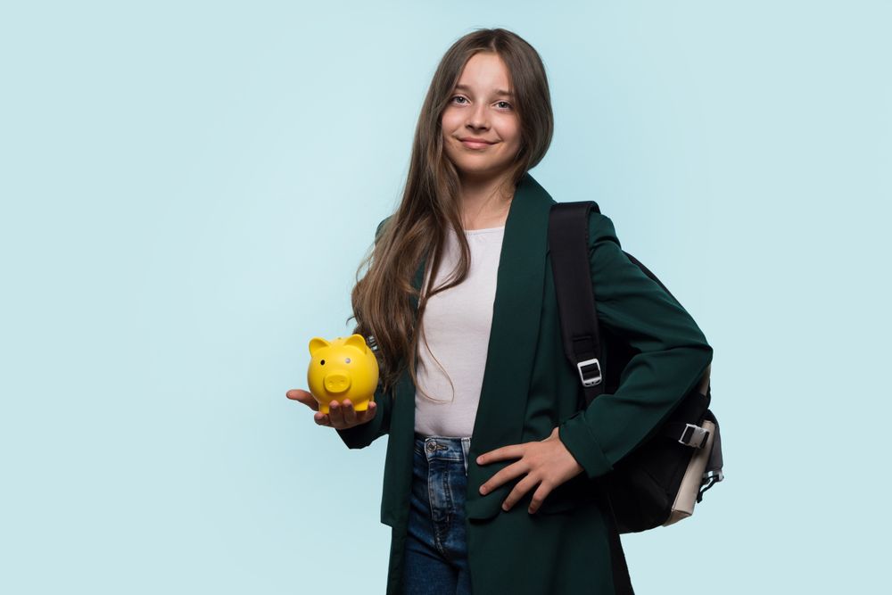 Teen girl holding yellow piggy bank, wearing backpack, smiling confidently.