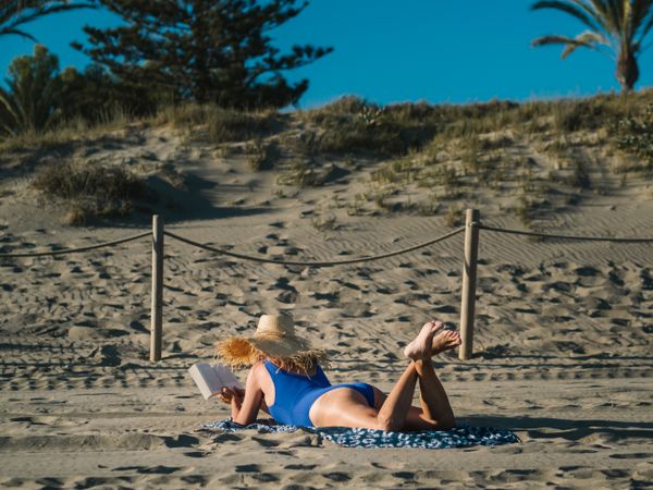 Sandy beach and woman in big hat lying and reading book