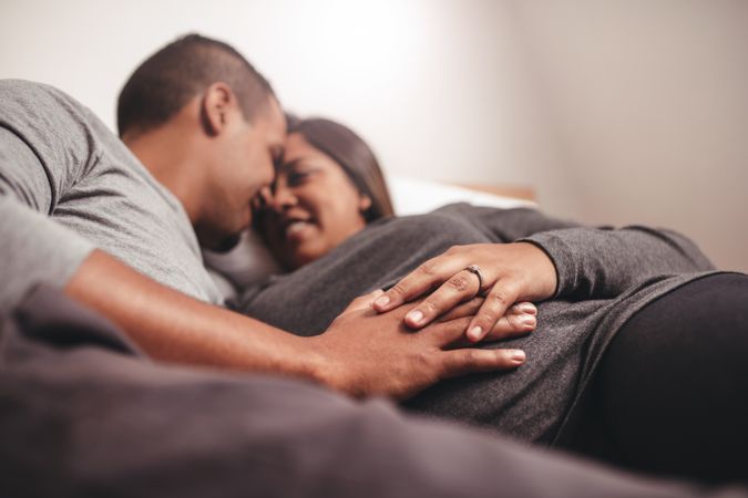 Close up of happy couple lying on bed holding hands over expectant mother’s belly