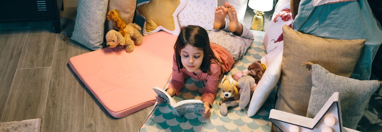 Banner of serene little girl looking a book while resting surrounded by cushions in homemade teepee