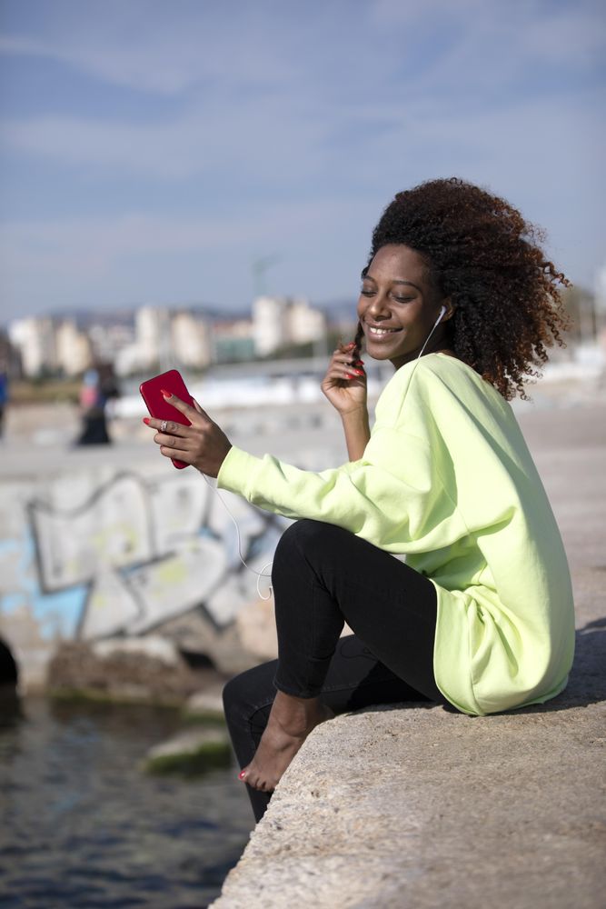 Smiling female with curly hair in bright green shirt sitting listening to music on dock