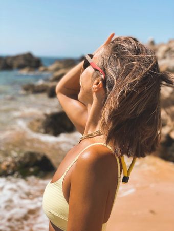 Side view of blonde woman in yellow bikini wearing sunglasses standing on beach