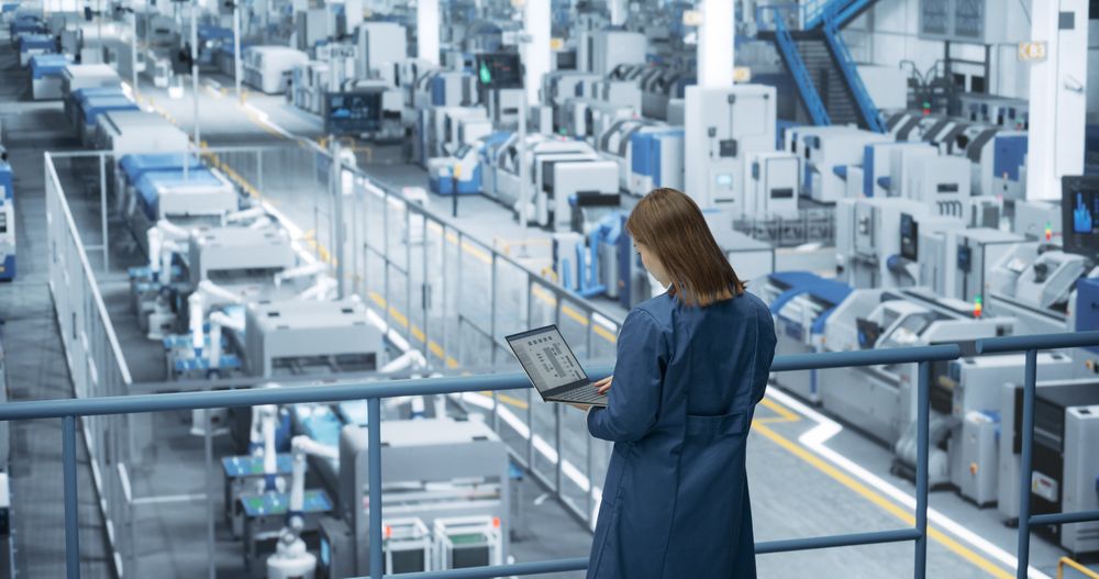 Asian Female Engineer Standing in a Factory Facility, Working on Laptop Computer, Researching and Developing Technologies to Produce Electronics at a Modern Automated Manufacture with Robotic Arms