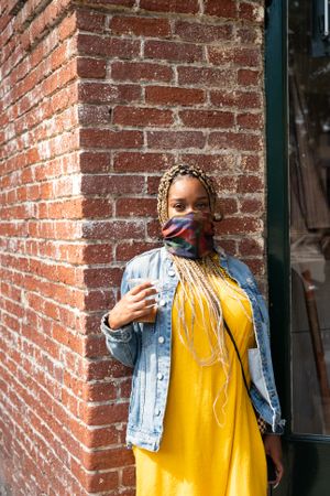 Woman wearing neck gator for COVID protection leaning against brick wall outside