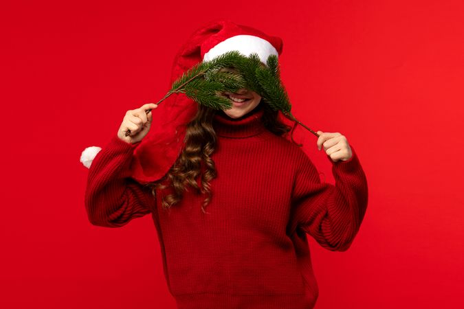 Girl in Santa hat holding fir branches over face.