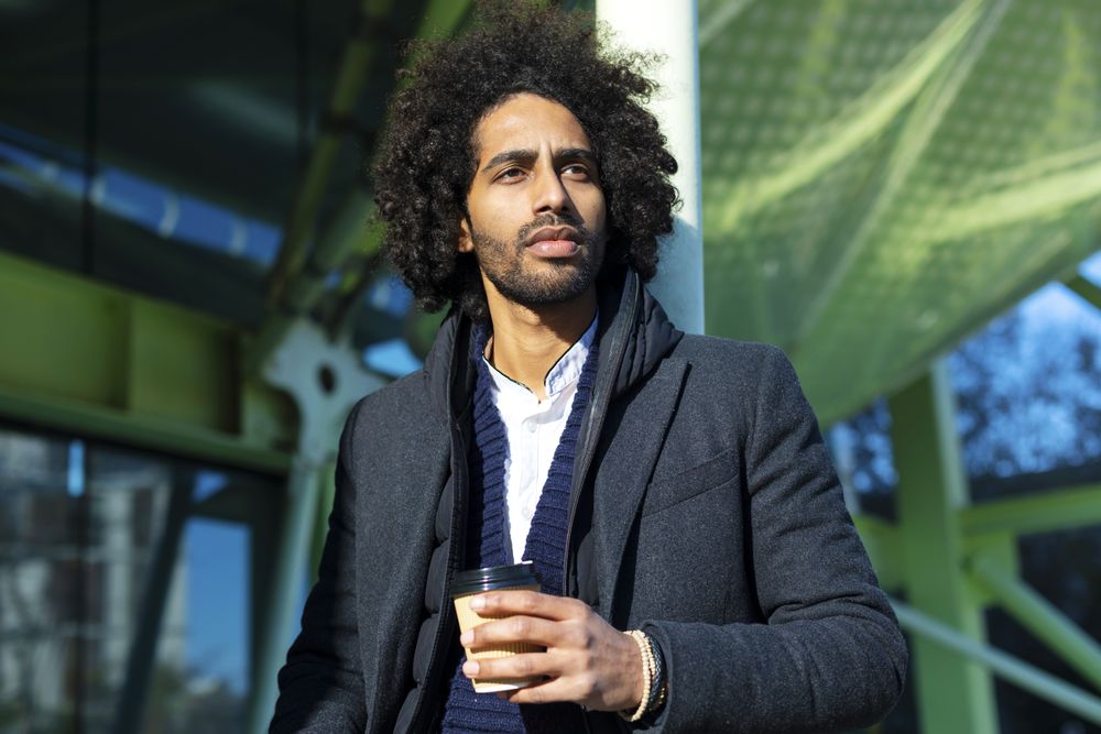 Portrait of Black man in jacket standing outside of building holding warm beverage while looking in the distance