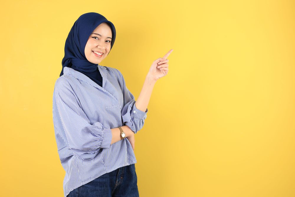 Smiling Beautiful Asian Young Woman in Blue Striped Shirt and Navy Jilbab Pointing Fingers Aside