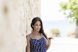Latina woman in blue dotted dress leaning on wall outside and looking away playing with hair