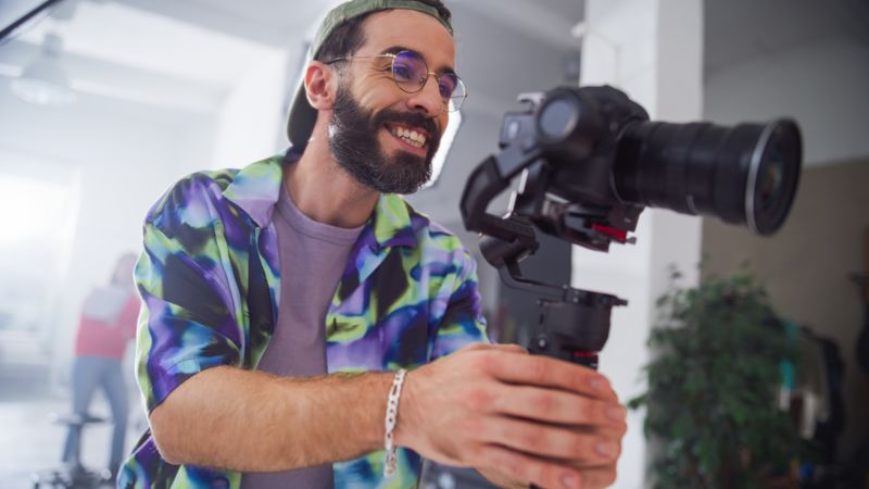 Young Male Videographer With Beard, Wearing A Colorful Jacket, Operates A Professional Camera In A Well-lit Studio, Capturing Engaging Content With A Focused And Joyful Expression.