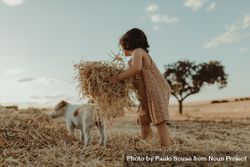 Little girl lifting a pile of hay with a dog in the background bxVRd5