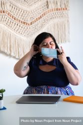 Woman sitting in a modern office with her laptop, adjusting her face mask 0WVlWb