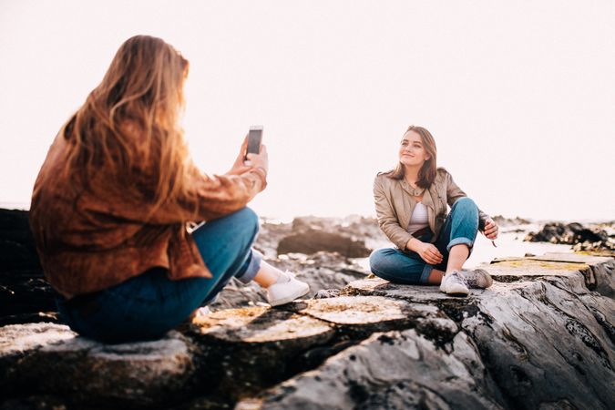 A young woman taking a friend’s photo sitting atop rocks