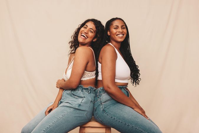 Two cheerful female friends sitting together on a wooden chair while wearing denim jeans and bras