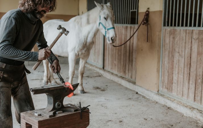 Blacksmith forging horseshoe near stable