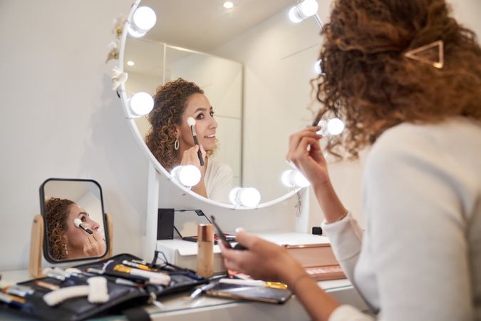 Positive woman applying cosmetic with brush on face looking in m