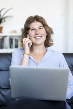 Businesswoman sitting on sofa at home using a laptop and speaking on phone
