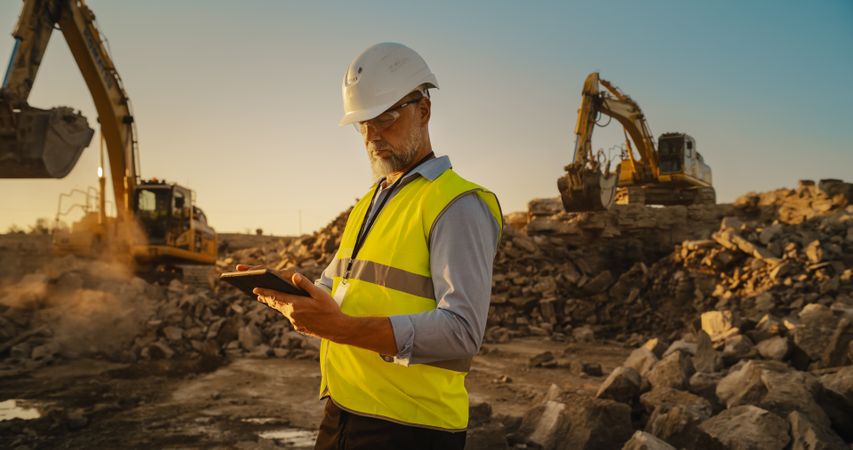 Male Civil Engineer Wearing Protective Goggles And Using Tablet On Construction Site On A Sunny Day. Man Inspecting Building Progress. Excavator Loading Materials Into Big Industrial Truck.