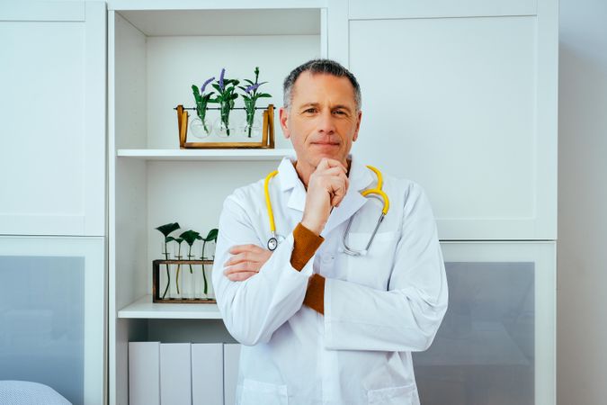 Thoughtful mature doctor standing in a clinical setting with hand to his chin