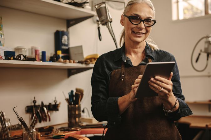 Portrait of happy mature woman jewelry maker using digital tablet in her workshop