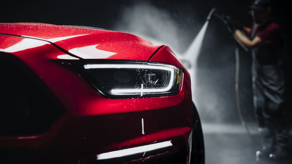 Advertising Style Photo of a Professional Car Wash Specialist Using a High Pressure Washer to Clean and Prepare a Red Sports Coupe for Detailing