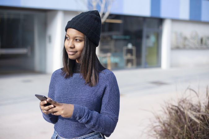 Female in wool hat and blue sweater checking smart phone outside on street with space for text