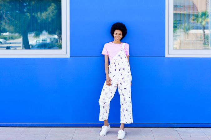 Smiling woman in floral coveralls leaning against outside of blue building