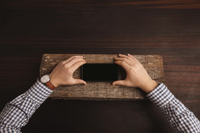 Man wearing plaid shirt and leather watch holding cell phone on slab of wood on table