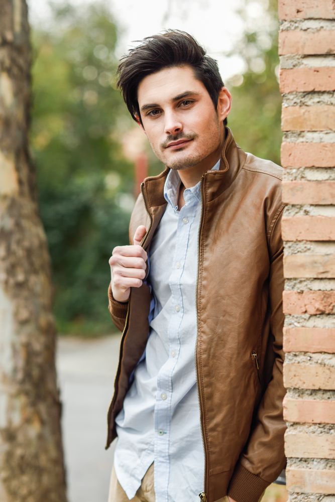 Man in leather coat leaning on brick wall outside