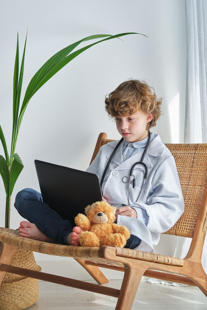 Boy doctor with toy bear and laptop on wicker chair