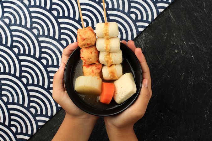 Hands holding bowl of of Japanese oden, skewers of dumplings and seafood
