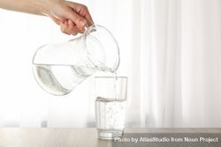 Hand pouring water into glass from pitcher in bright room near window 4ZYxy5