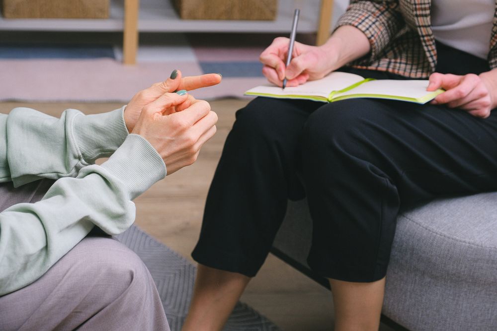 Cropped image of a person in sweater sitting on couch talking to a person writing on a notebook
