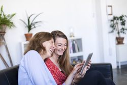 Two laughing females at home sitting on sofa using digital tablet
