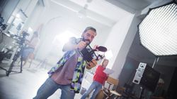 Focused Young Male Cinematographer With Beard, Wearing A Cap And Plaid Shirt, Actively Filming In A Professional Studio Setting, Surrounded By Modern Filming Equipment And Crew Members.