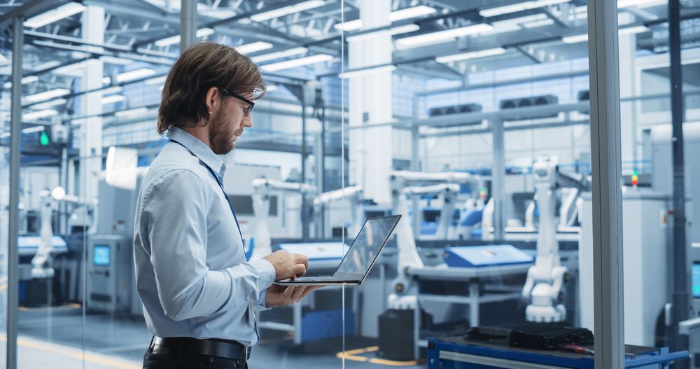 Factory Manager Working on Laptop at Futuristic Industrial Manufacture with Autonomous Support Robots. Specialist Monitoring Technical Data on a Computer, Analyzing Reports and Planning Updates