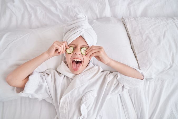Boy lying on bed and putting cucumber slices on eyes